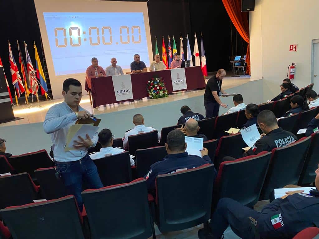 Personas sentadas en un auditorio y una entregándoles una hoja, una mesa, banderas, paredes y una pantalla proyectora
