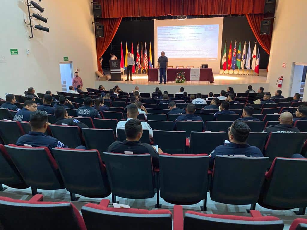 Personas sentadas en un auditorio, una mesa, banderas, paredes y una pantalla proyectora