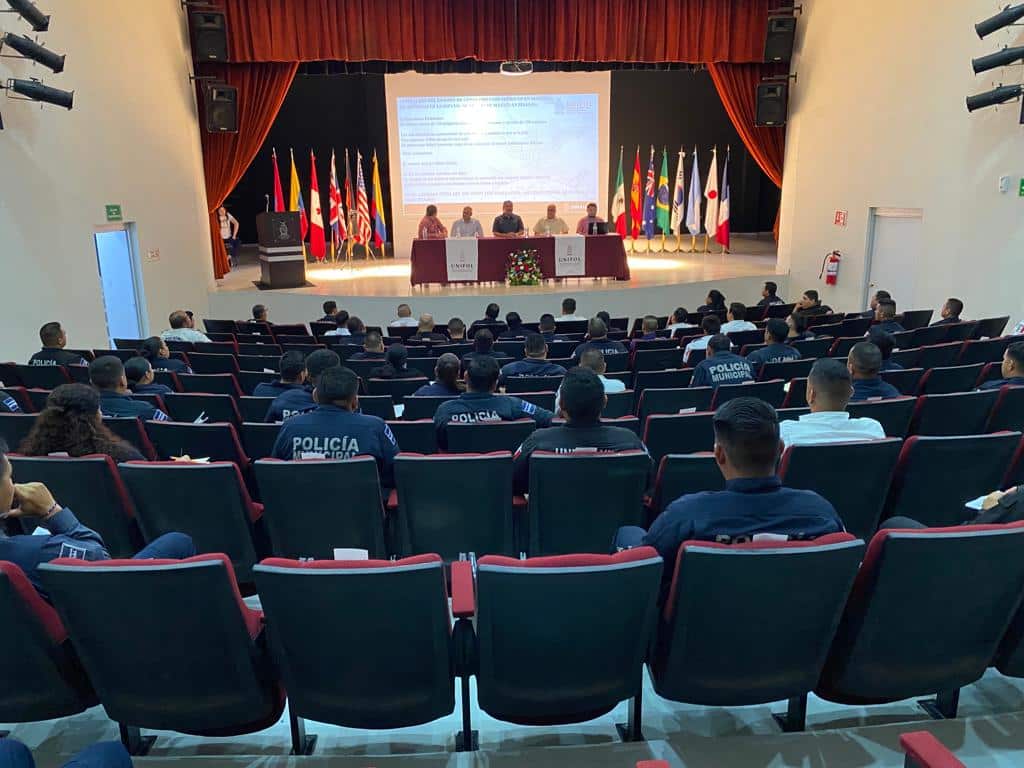 Personas sentadas en un auditorio, una mesa, banderas, paredes y una pantalla proyectora