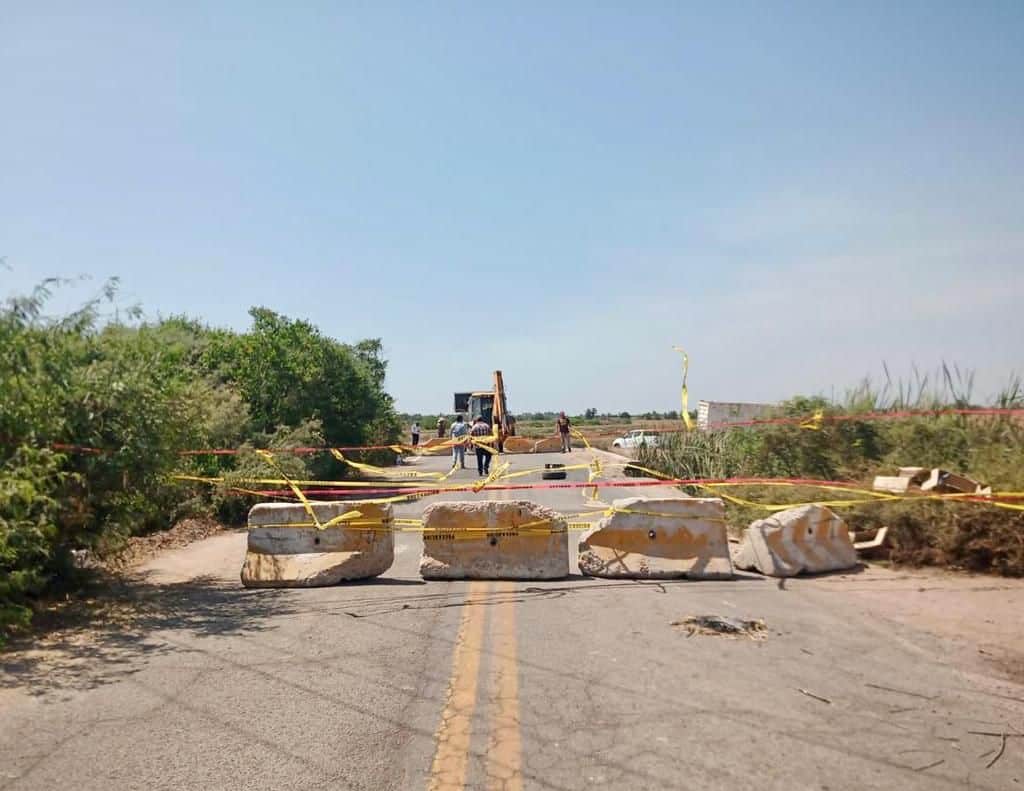 Bloques de guarnición obstruyendo el paso de la carretera, cintas de color amarillo y rojo, al fondo personas, una máquina y una camioneta