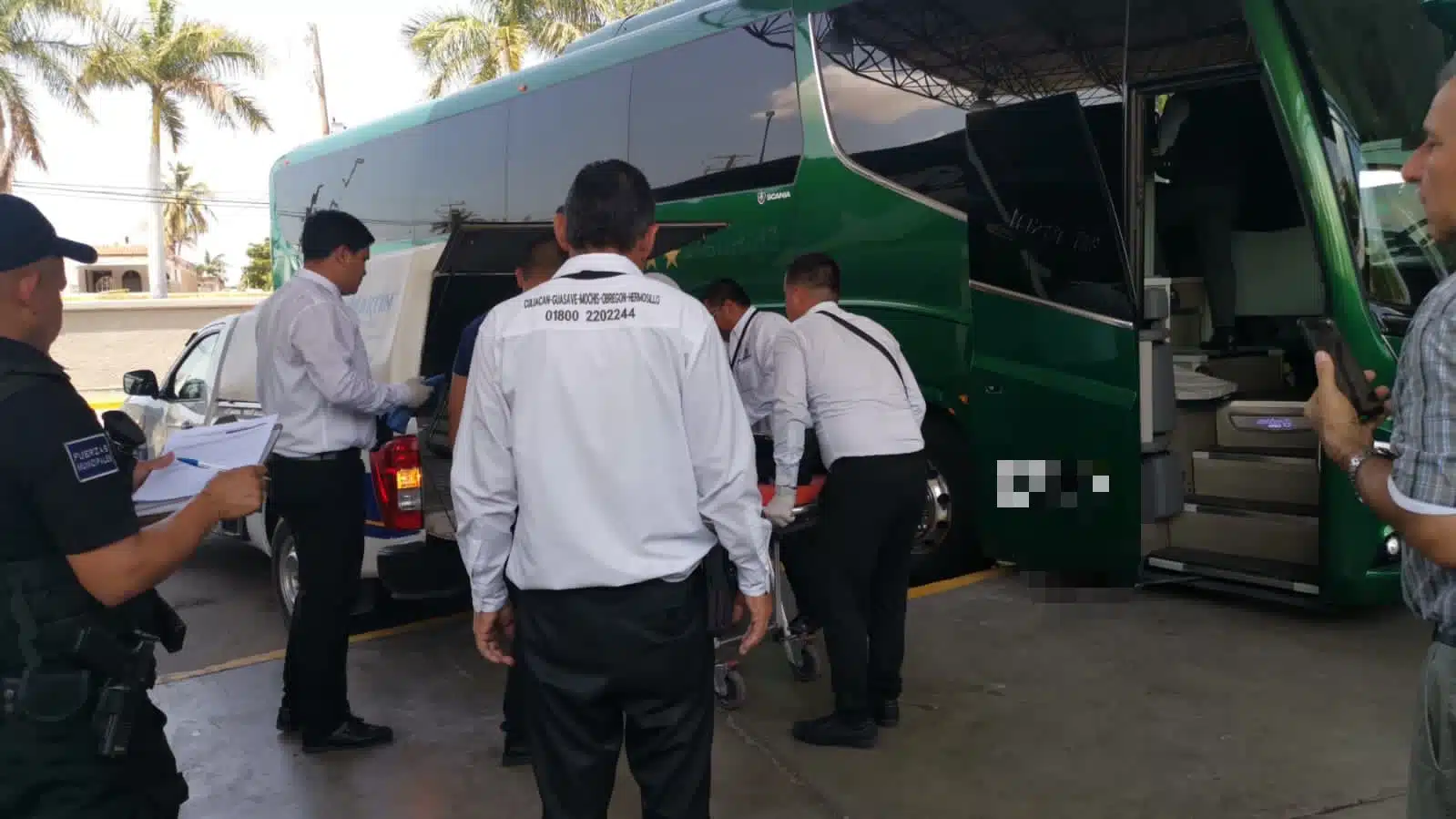 2 personas moviendo una tipo camilla y 3 más mirando, una camioneta funeraria y la parte de un autobús de pasajeros