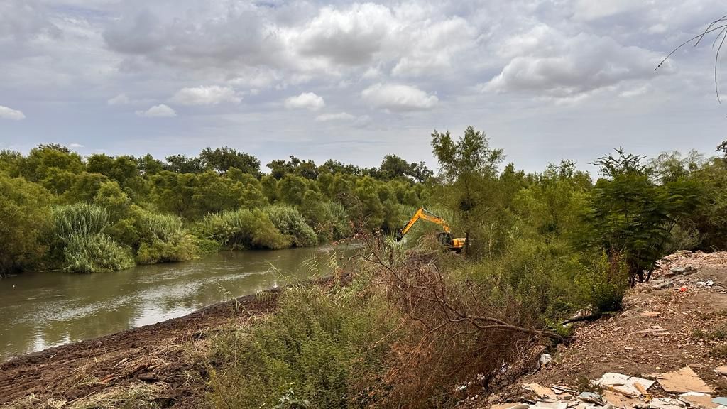 Una retroexcavadora a un lado del río, agua, árboles, tierra y cielo con nubes