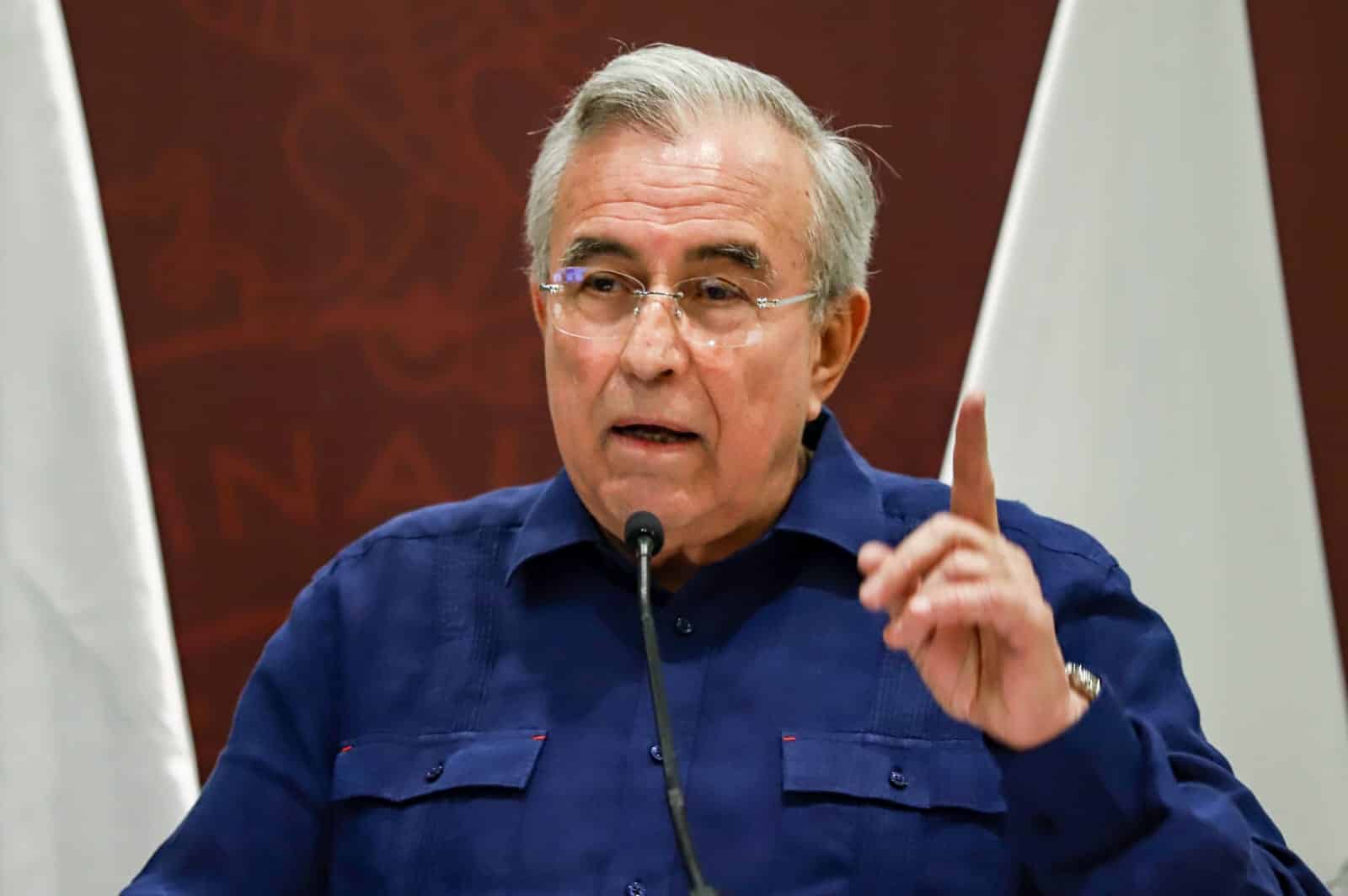 Una persona con lentes alzando su dedo indice y con un micrófono de podium