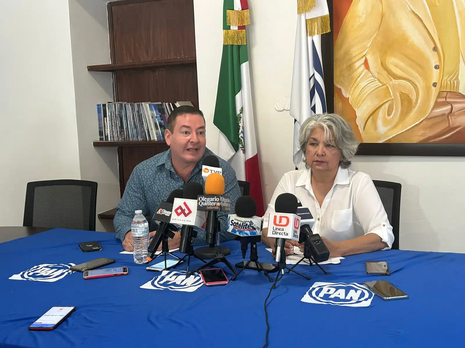 2 personas sentadas, micrófonos de medios de comunicación, sillas, cuadros, paredes, 2 banderas y una mesa con mantel azul y letras blancas