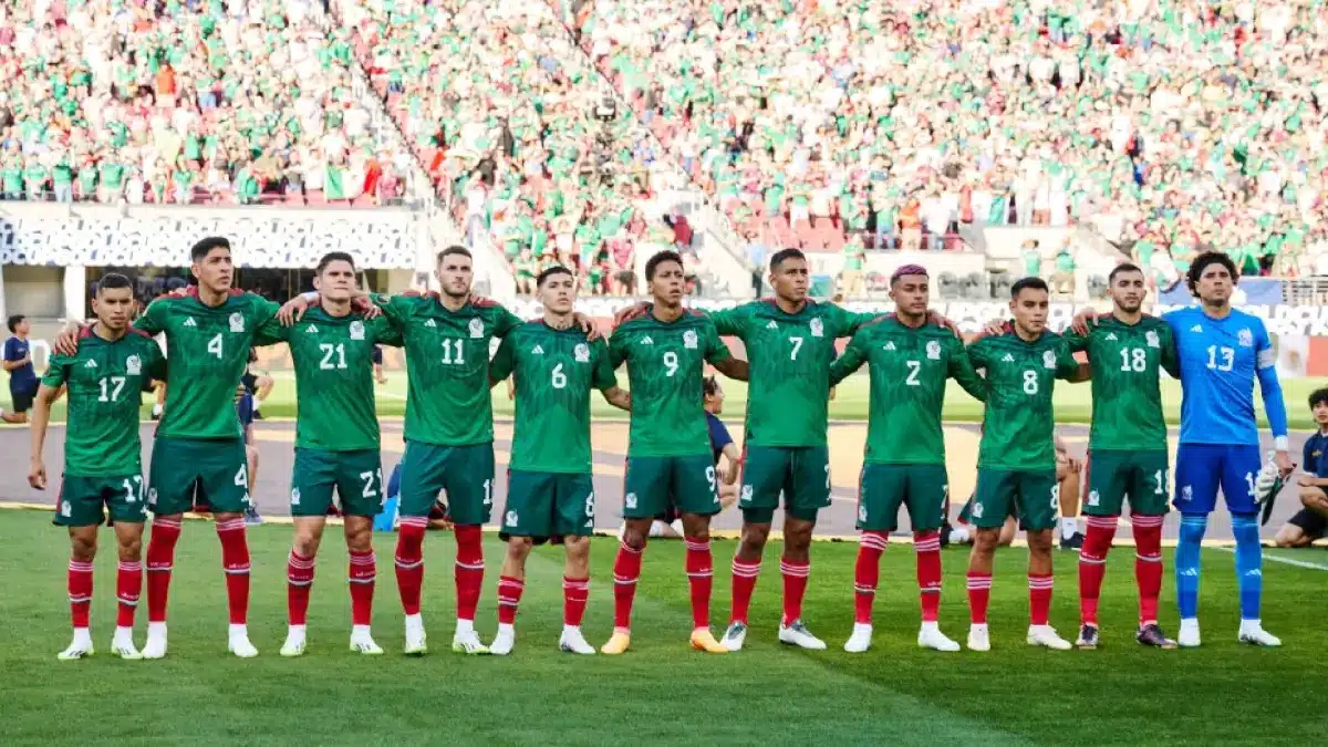 11 hombres abrazados parados sobre el césped de una cancha de futbol, 10 de ellos traen playera verde y uno de color azul, al fondo mucha gente en las gradas