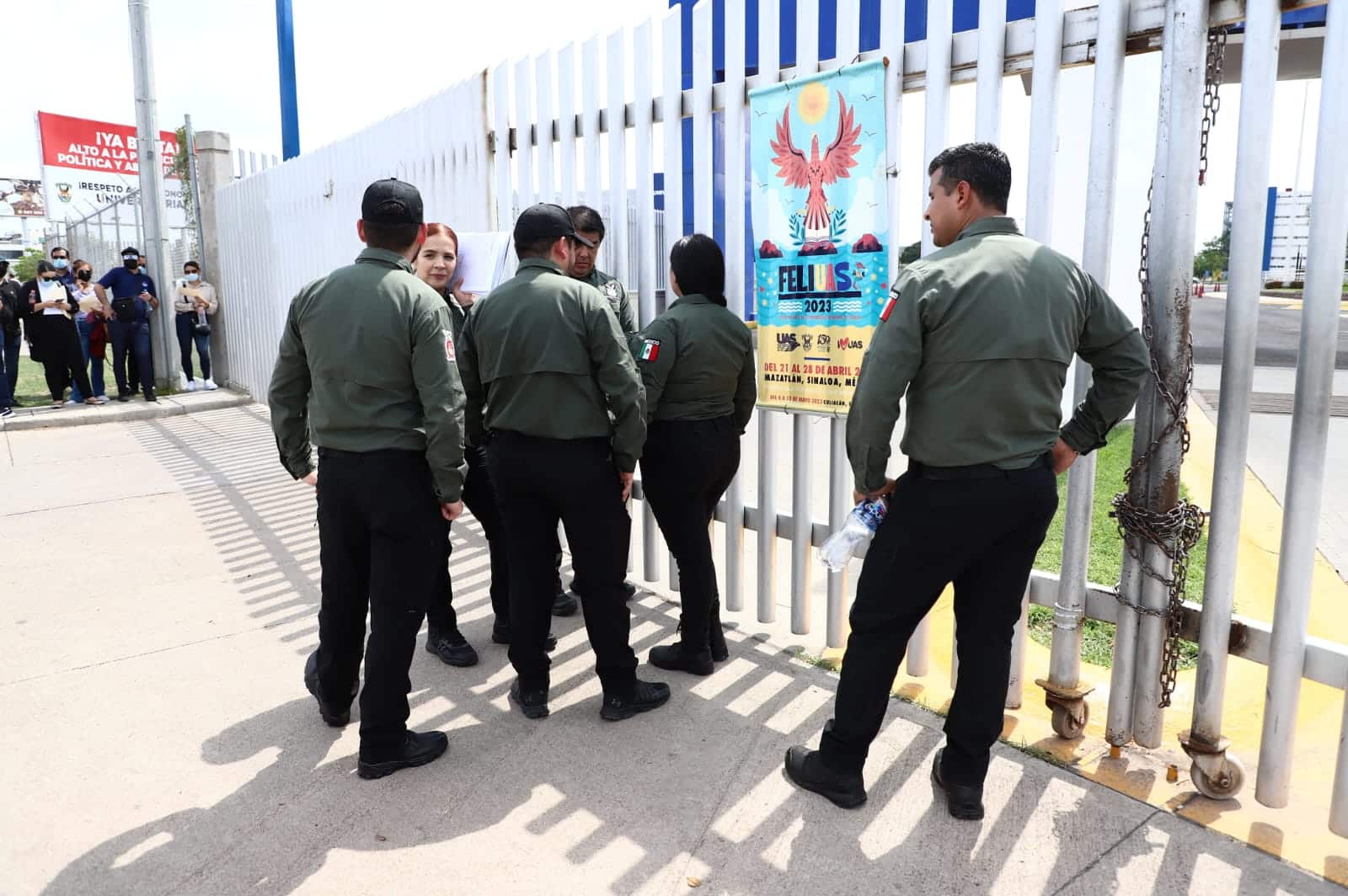 6 personas paradas a un lado de una reja de una escuela, un cartel y al fondo más personas