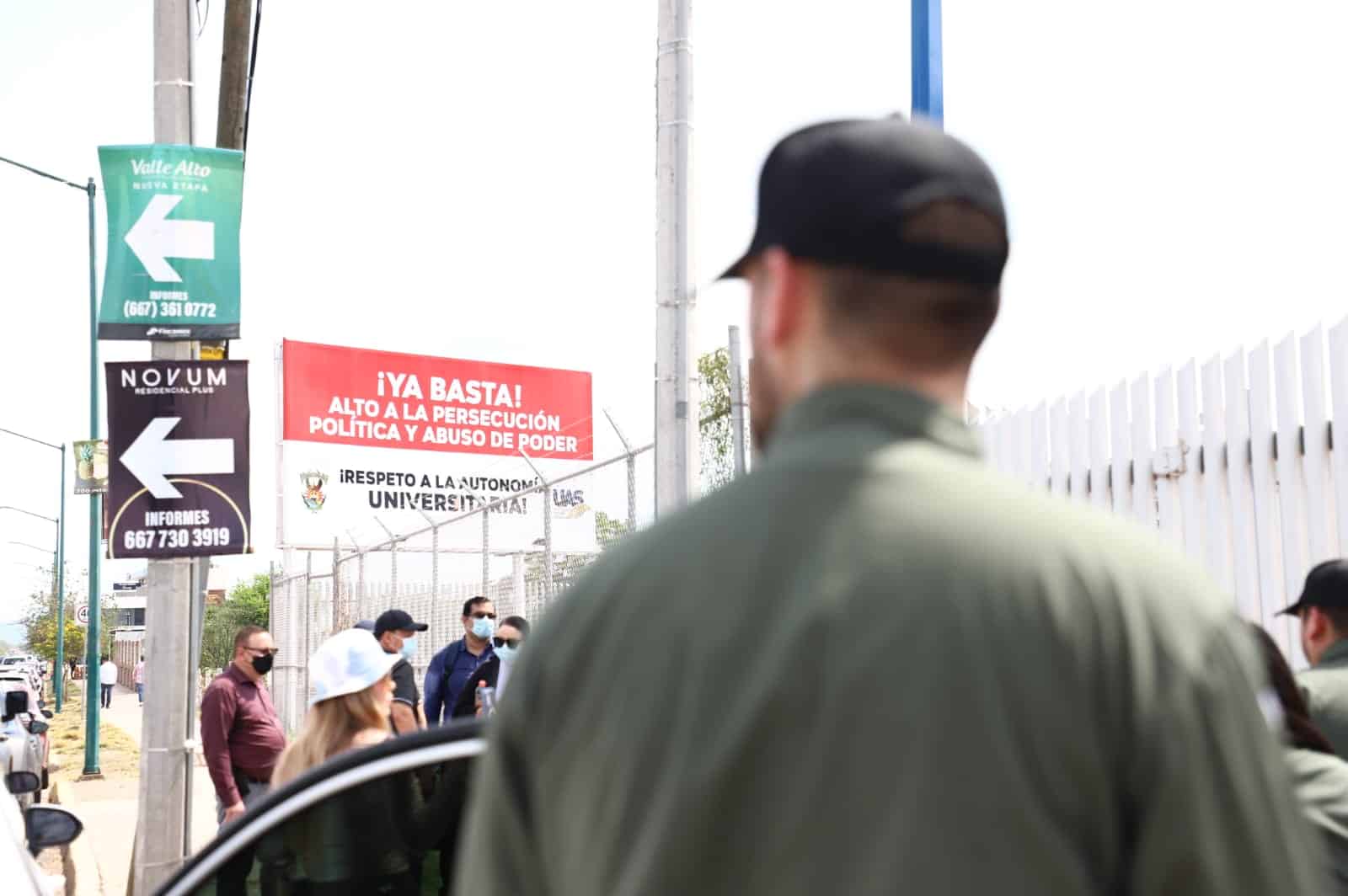Una persona con gorra de espaldas, personas, un poste, reja de una escuela, carteles y un poste