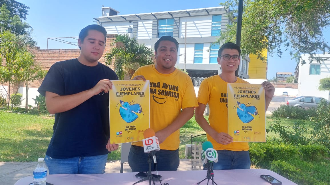 3 personas, 2 carteles, 2 micrófonos de medios de comunicación sobre una mesa color blanca, 2 celulares, una botella de agua, árboles, césped, un carro estacionado y casas al fondo