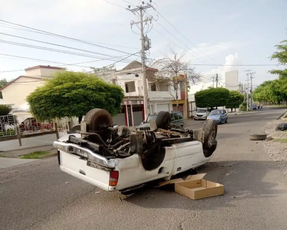 Camioneta con las llantas hacia arriba, casas, árboles, carros, postes y cables