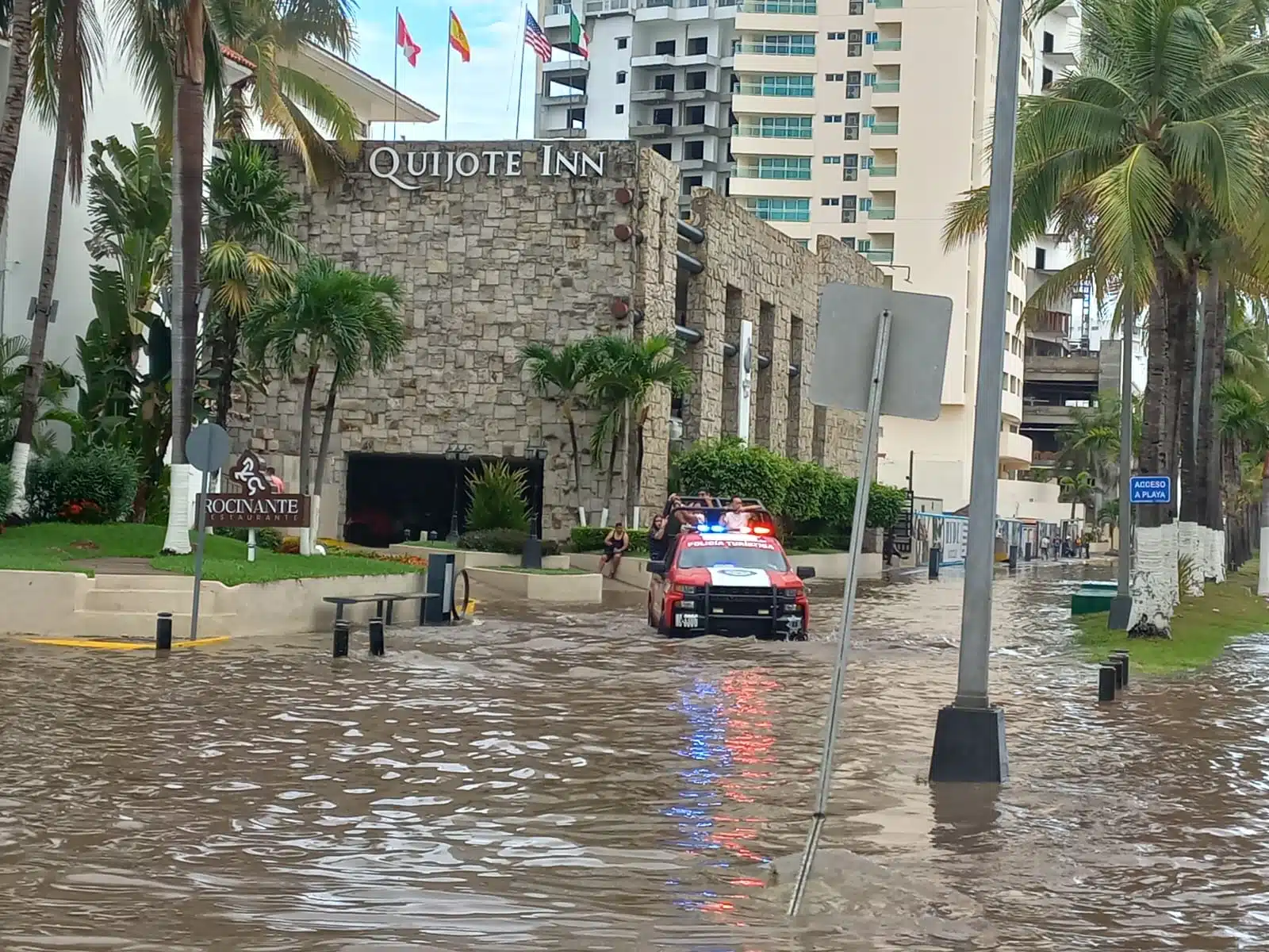Una camioneta con personas arriba que va pasando por una calle inundada, edificios, palmeras y postes al fondo