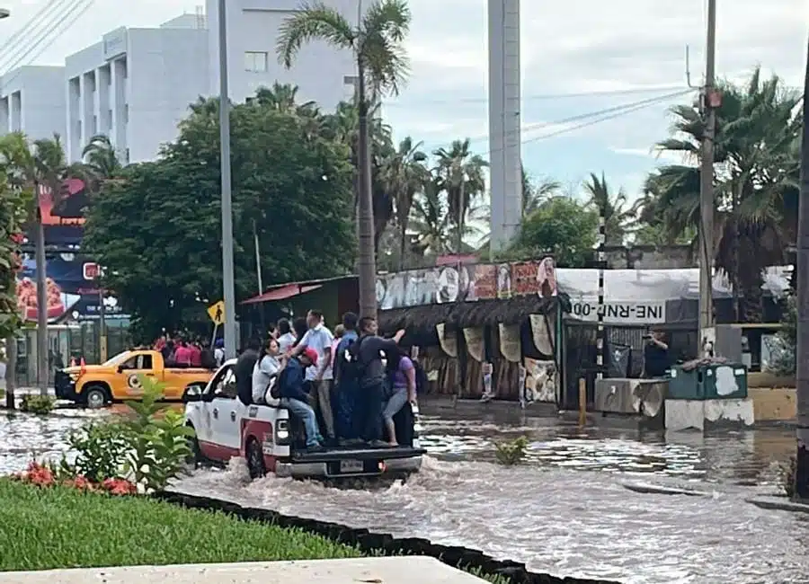 Personas arriba de una camioneta que va pasando por una calle inundada, edificios, palmeras y árboles al fondo