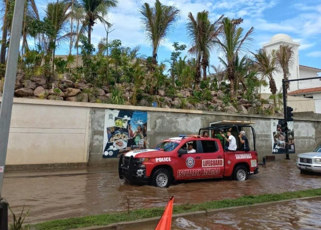Una camioneta con personas arriba que va pasando por una calle inundada, edificios, palmeras y una pared al fondo