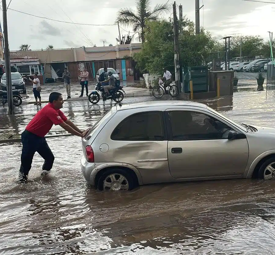 Una persona empujando un carro que quedó sobre una calle inundad, personas y carros al fondo