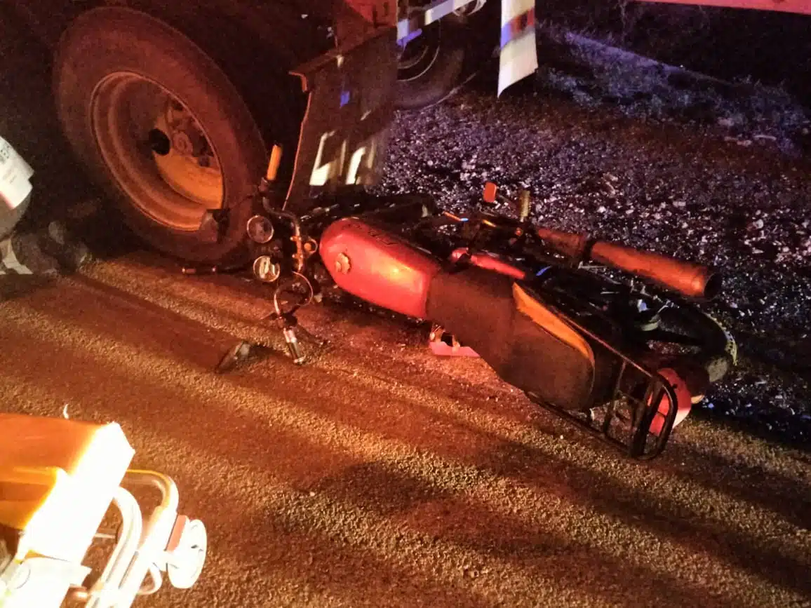 Motocicleta derribada sobre el pavimento al lado de la llanta trasera del tráiler