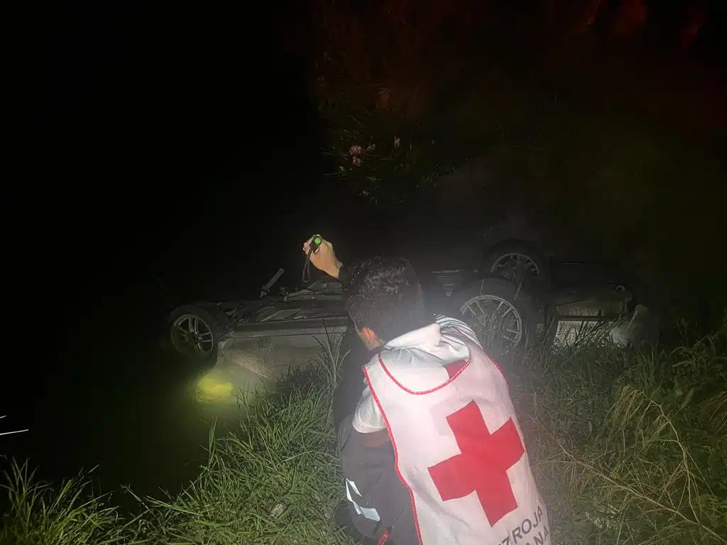 Una persona mirando y aluzando hacía el lugar del accidente, un carro con los neumáticos hacia arriba