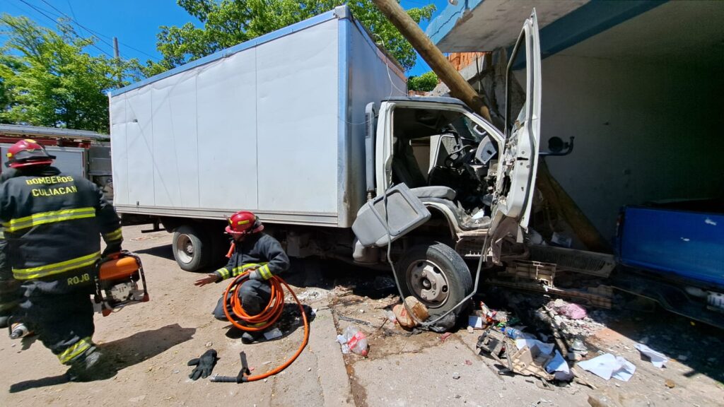 Un camión chocado de la parte de enfrente, 2 personas con vestimenta de bomberos con mangueras