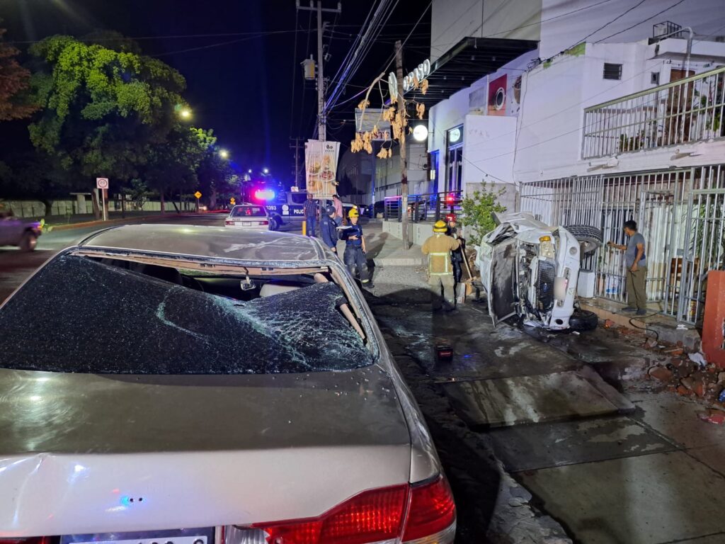 Personas mirando hacia una camioneta volcada sobre la banqueta, casas y carros estacionados