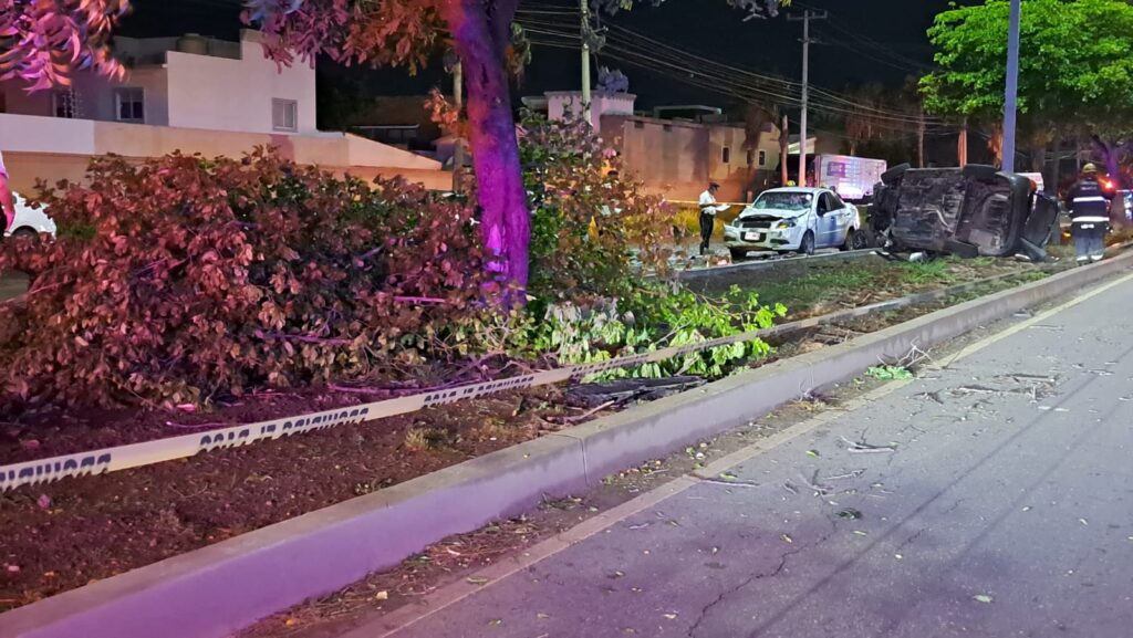 Un carro volcado y otro chocado del frente, camellón, árboles y cinta amarilla delimitando el área del accidente