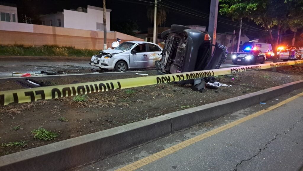 Un carro volcado y otro chocado del frente, camellón, árboles y cinta amarilla delimitando el área del accidente