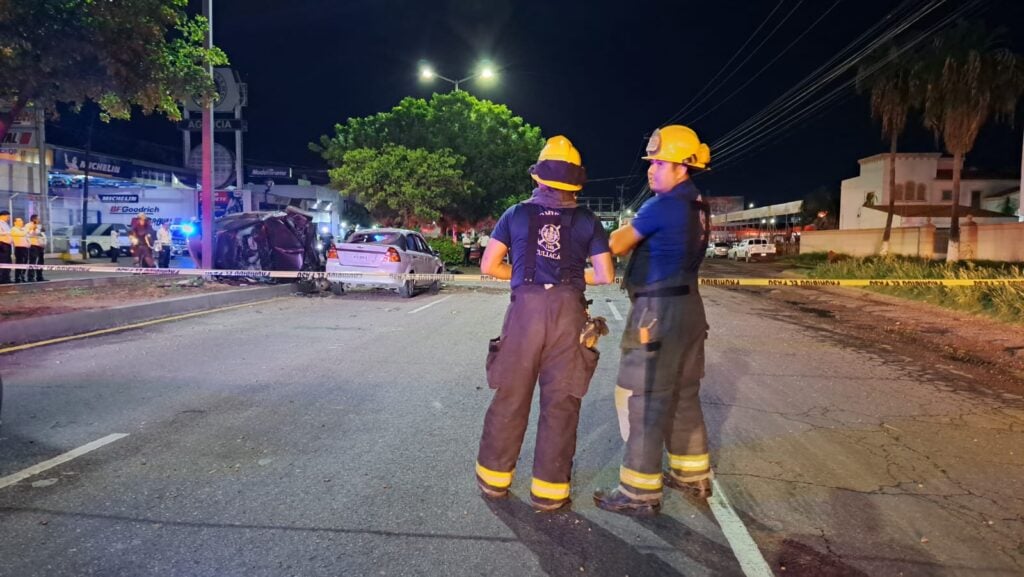 2 personas paradas con uniforme de bomberos uno mirando hacia el lugar del accidente y el otro hacia otro lado, un carro volcado sobre el camellón y otro sobre la carretera, árboles y cinta amarilla delimitando el área del accidente