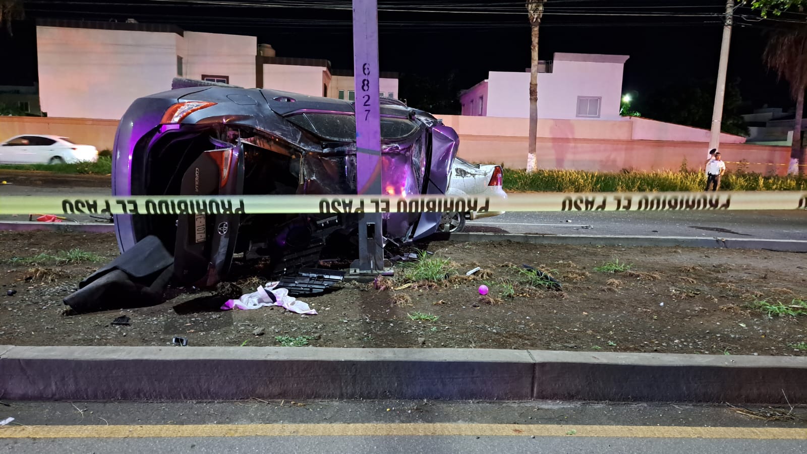 Un carro volcado sobre el camellón central, un poste, cinta amarilla delimitando el área de un accidente y al fondo casas de un fraccionamiento