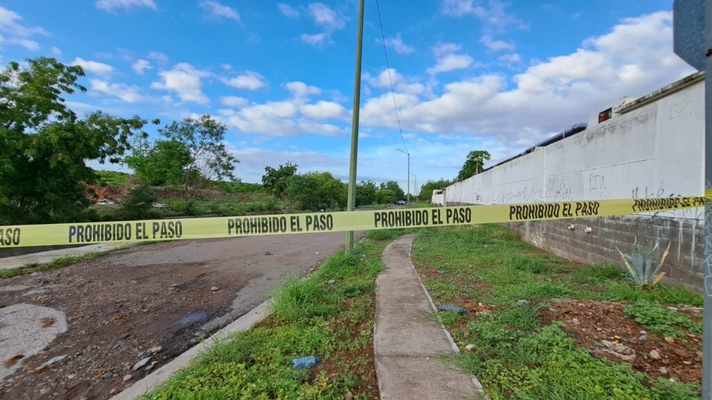 Una cinta amarilla delimitando el área de un crimen, una barda balnca, postes, cielo con nubes, un canal y árboles