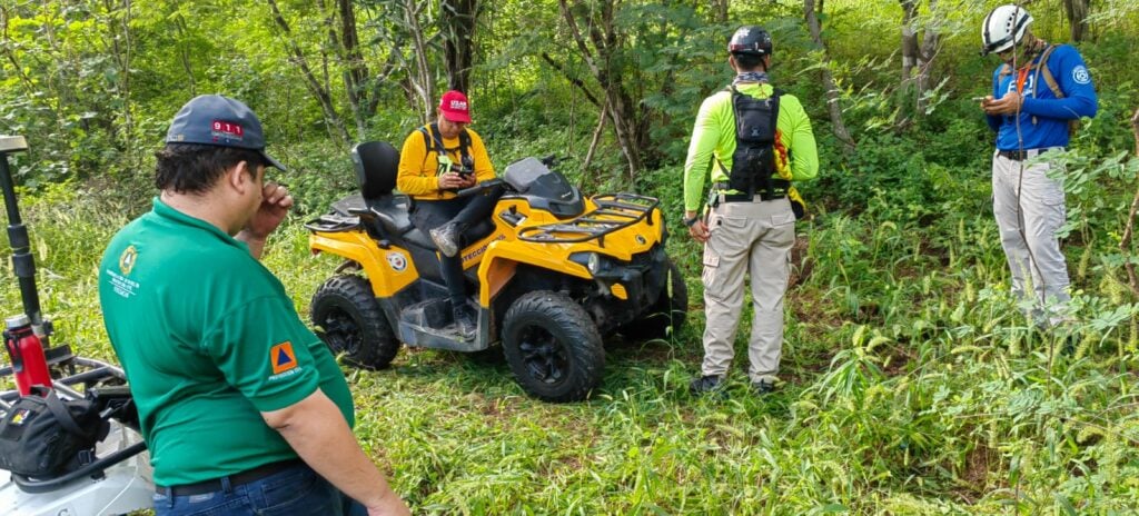 4 personas una sentada arriba de una cuatrimoto y las otras 3 de pie, árboles y plantas