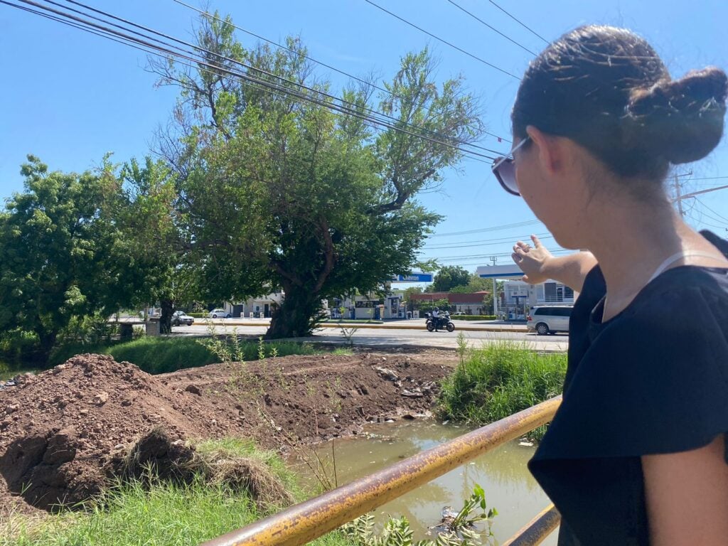 Una persona mirando a un canal con agua, árboles, cables y un tubo