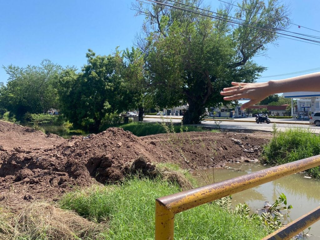 La mano de una persona, un canal com agua, un tubo, árboles y cables