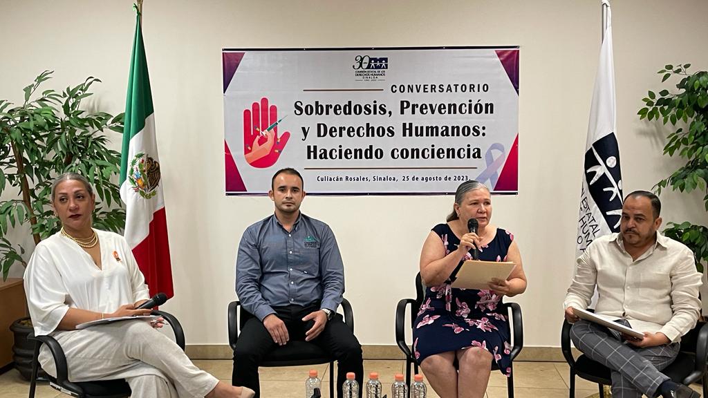 4 personas sentadas, 2 banderas y un cartel al fondo