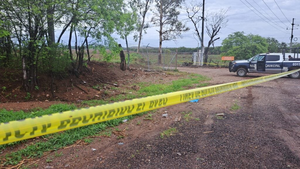 Cinta amarilla delimitando la escena de un crimen, árboles, terracería y una camioneta de la policía