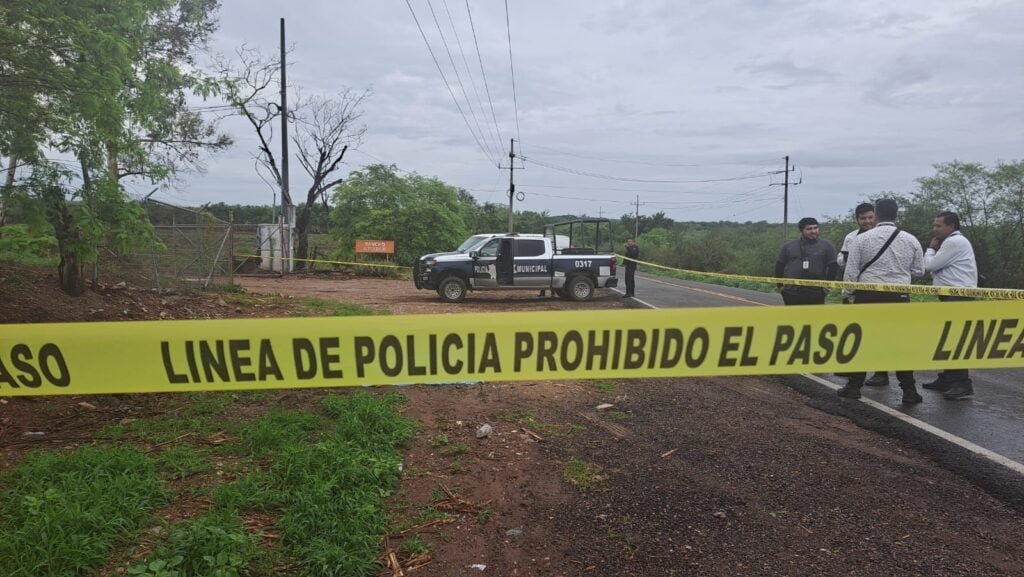 Cinta amarilla delimitando la escena de un crimen, árboles, terracería, personas y una camioneta de la policía