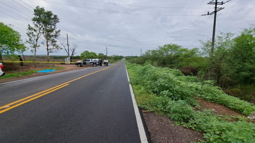 Cinta amarilla delimitando la escena de un crimen, árboles, terracería, personas, carretera y una camioneta de la policía