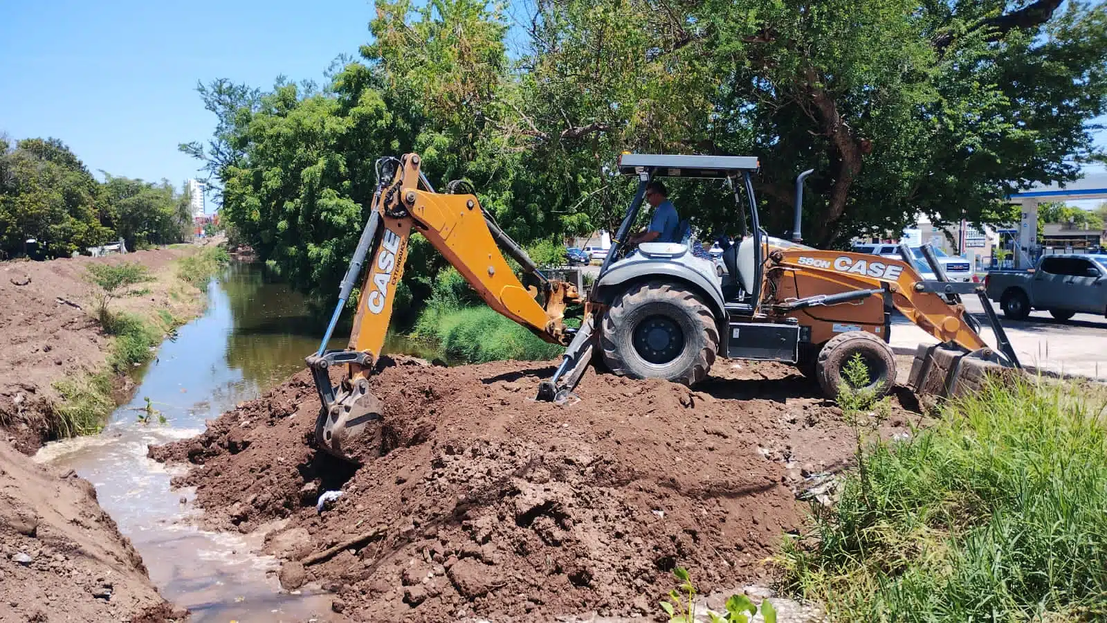 Una maquina retroexcavadora con tierra rellenando un canal con agua, árboles al fondo