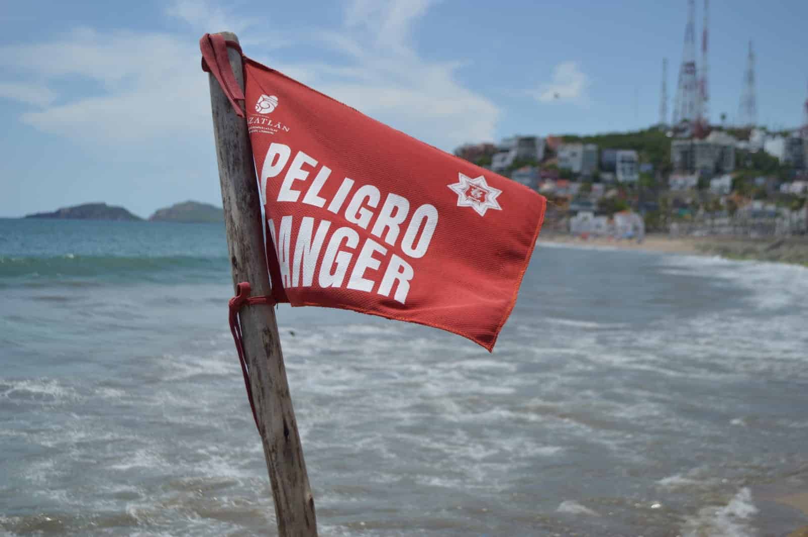 Bandera roja en la playa, agua del mar y al fondo un cero con casas