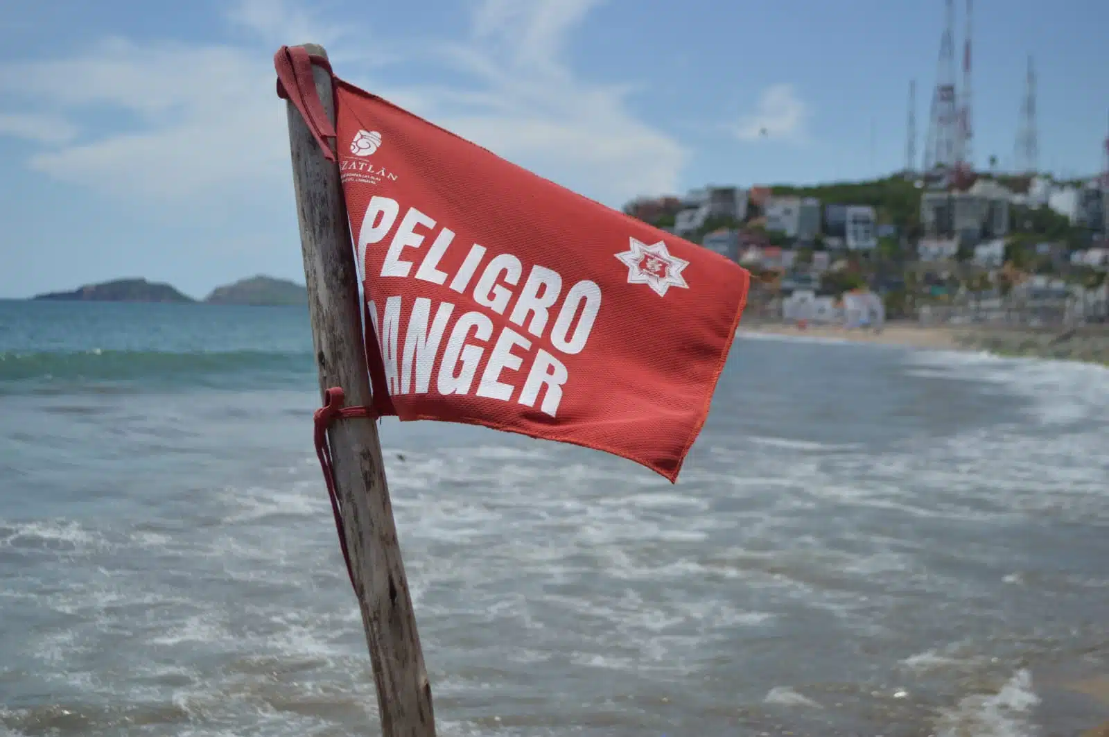 Bandera roja en la playa, agua del mar y al fondo un cero con casas
