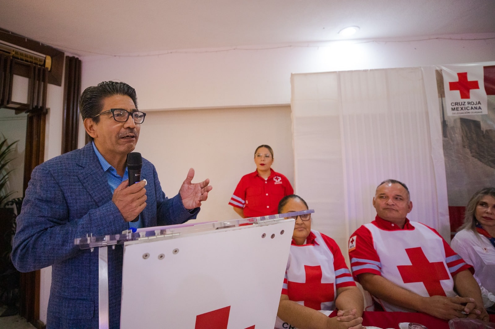 Una persona con lentes en un podium hablando y otras sentadas con uniforme de Cruz Roja