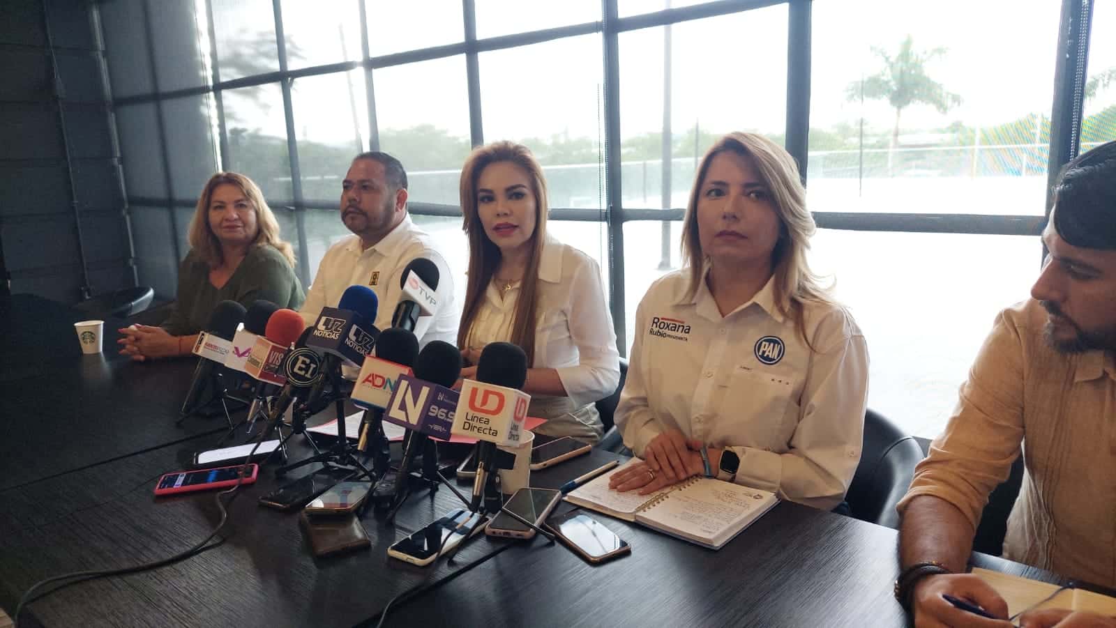 5 personas sentadas, una mesa y sobre ella una libreta, una carpeta, micrófonos, grabadoras y celulares de los medios de comunicación