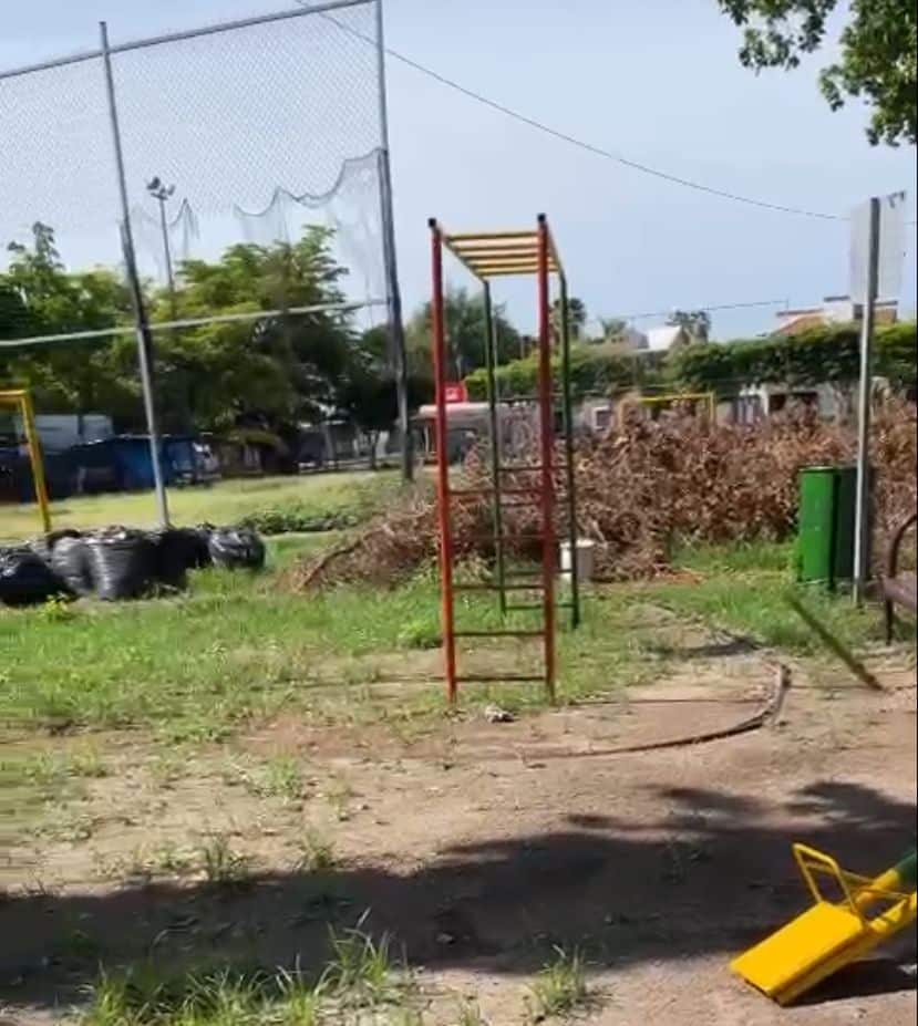 Un parque con juegos, árboles, tambos de basura, ramas y basura