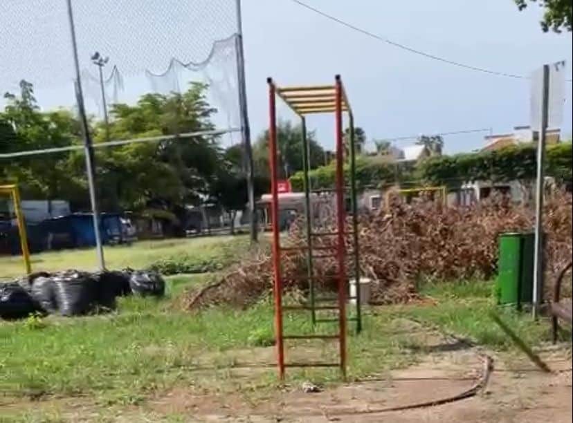 Un parque con juegos, árboles, tambos de basura, ramas y basura