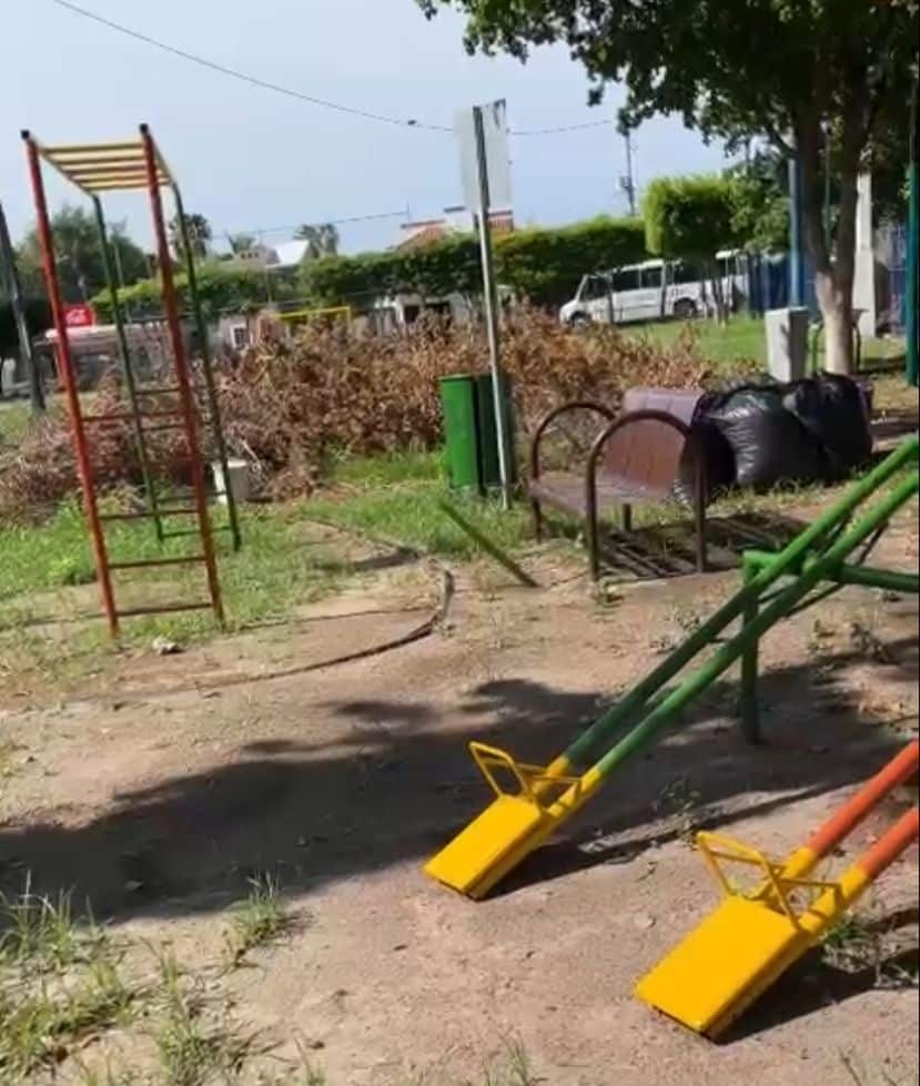 Un parque con juegos, árboles, tambos de basura, ramas y basura