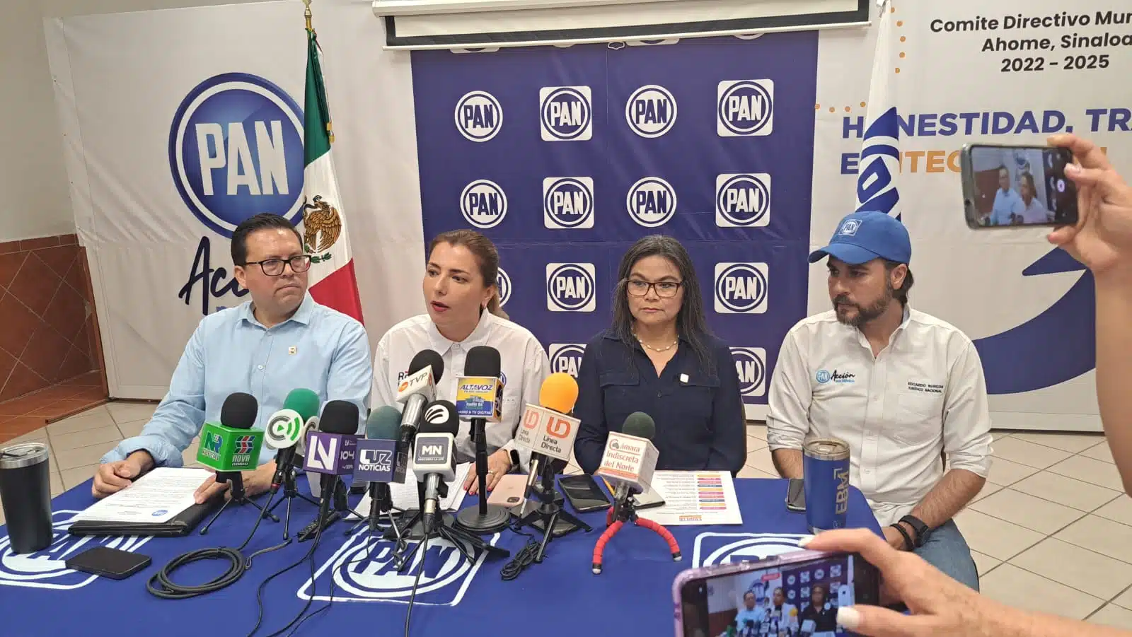 4 personas sentadas, una mesa con mantel azul y micrófonos de medios de comunicación