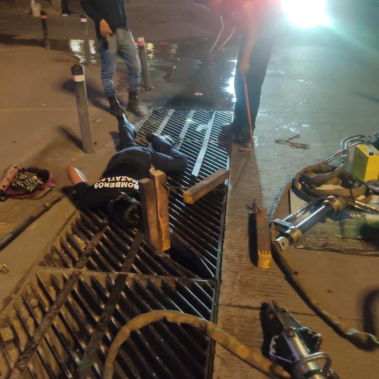 Una persona acostada metiendo la mano en una alcantarilla y herramientas de trabajo al lado