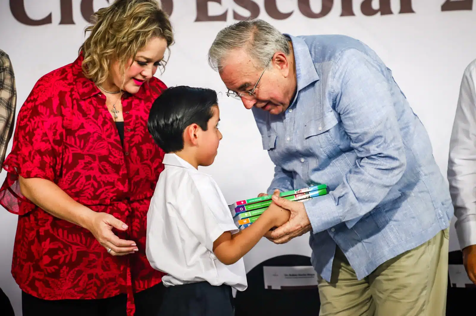 2 personas adultas entregándole libros escolares a un niño