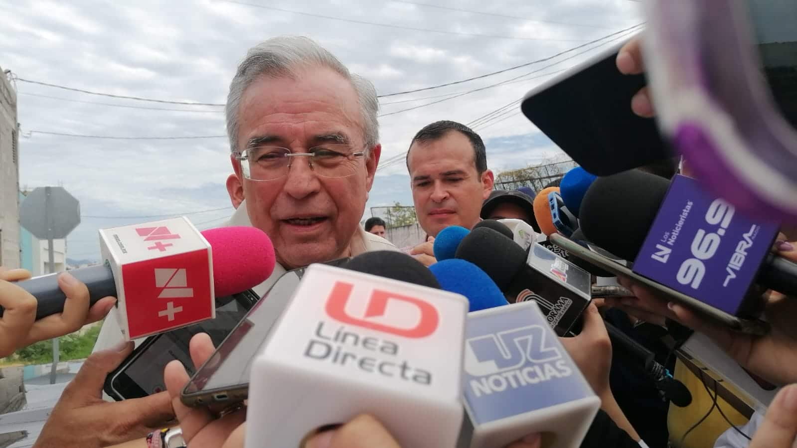 Una persona hablando, otra atrás de ella, y micrófonos de medios de comunicación