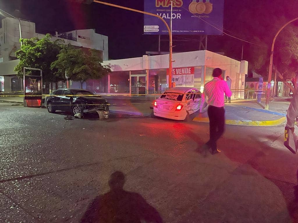Un taxi y un carro chocados, postes de los semáforos, árboles, negocios y una cinta amarilla delimitando el área del accidente