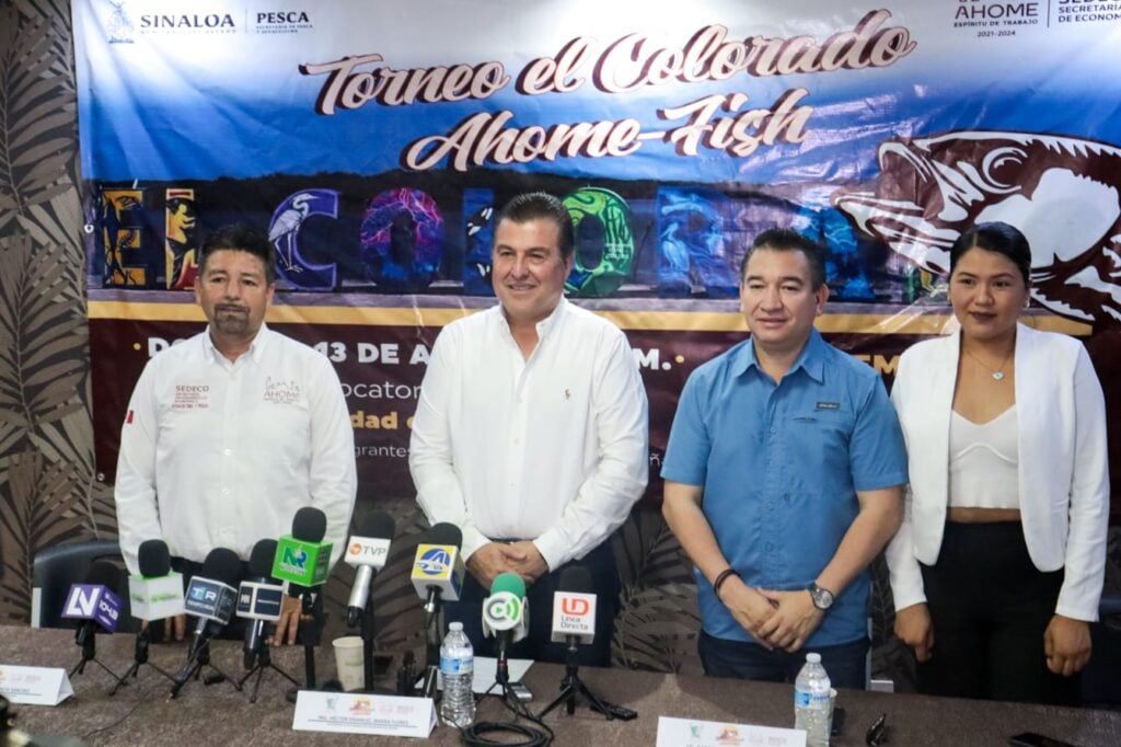 4 personas de pie posando para una foto, una lona al fondo y micrófonos de medios de comunicación sobre una mesa