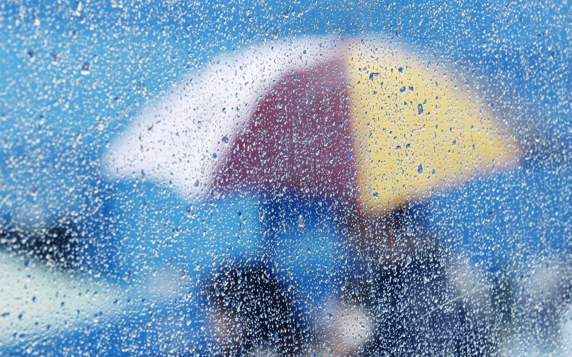 Gotas de lluvia sobre ventanal. De fondo hay personas con un paraguas colorido