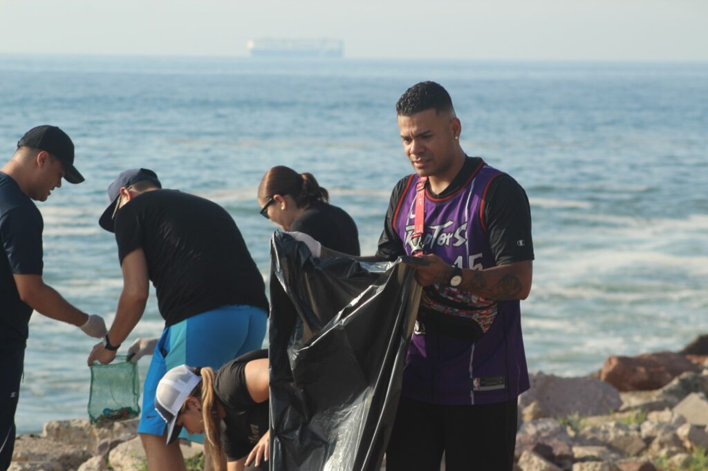 Joven con una bolsa de basura, al fondo gente recogiendo desechos y basura