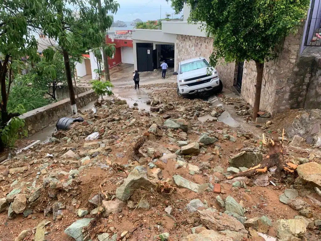 Piedras, tierra y escombros sobre una calle, carros, árboles y casas